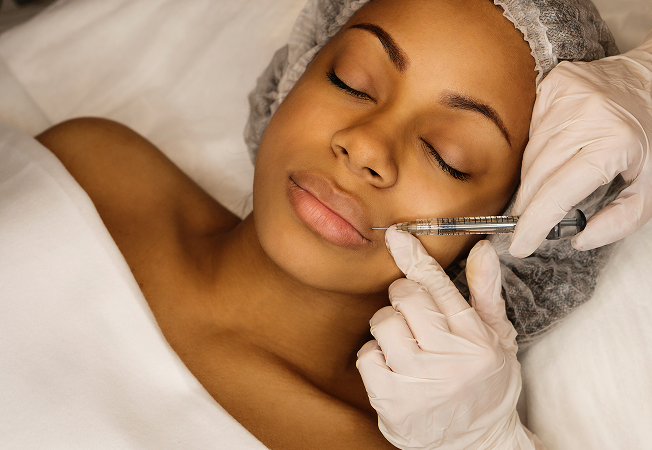 Woman with eyes closed receiving a cosmetic facial injection near her cheek from a gloved clinician.