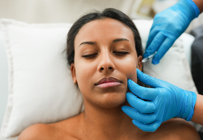 Woman with eyes closed as a clinician in blue gloves administers a facial injection near her jaw while she lies on a pillow-like support.